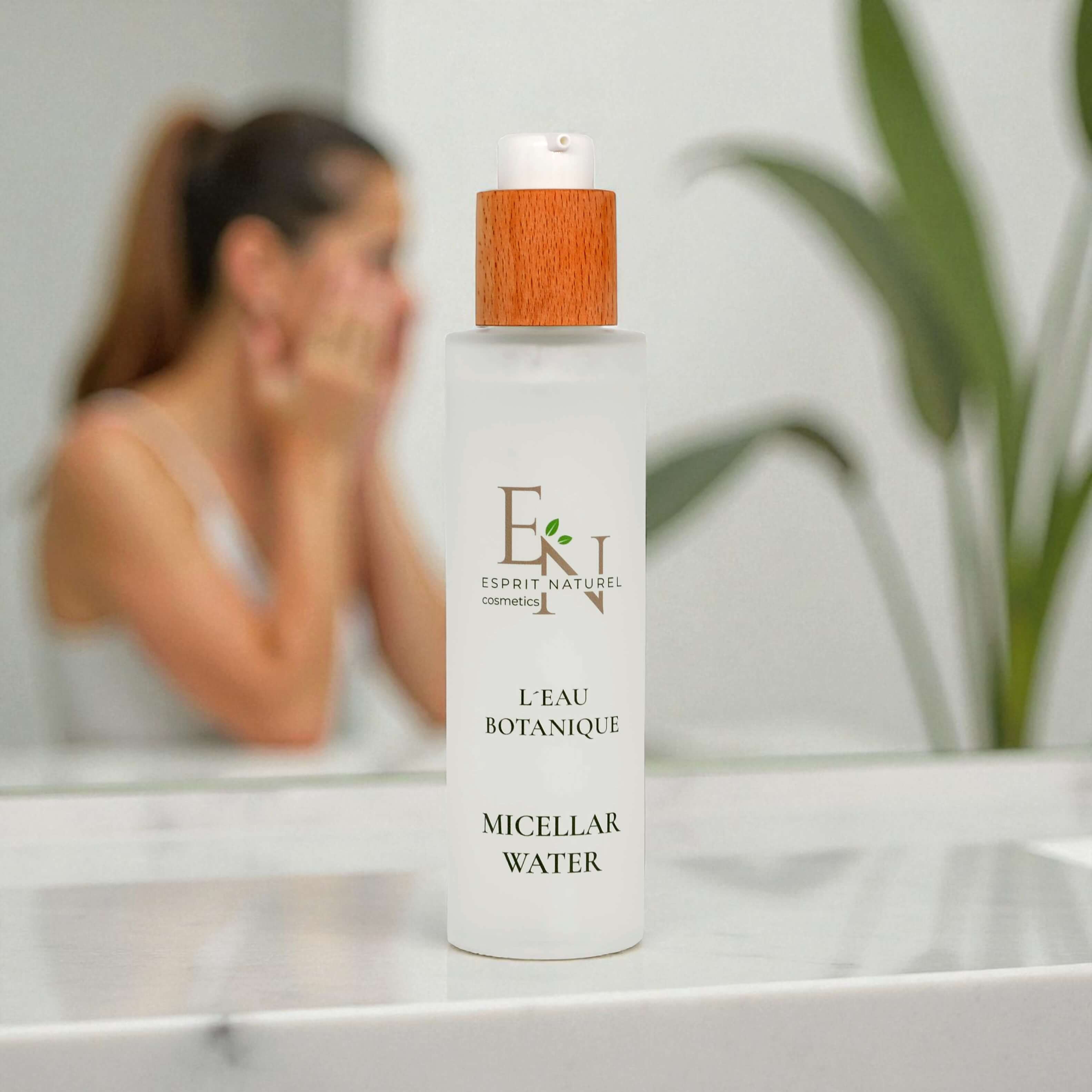 Esprit Naturel L’Eau Botanique Micellar Water auf dem Badezimmerwaschbecken mit Frau im Hintergrund, die sich das Gesicht reinigt – natürliche, vegane Gesichtspflege für reine und frische Haut.